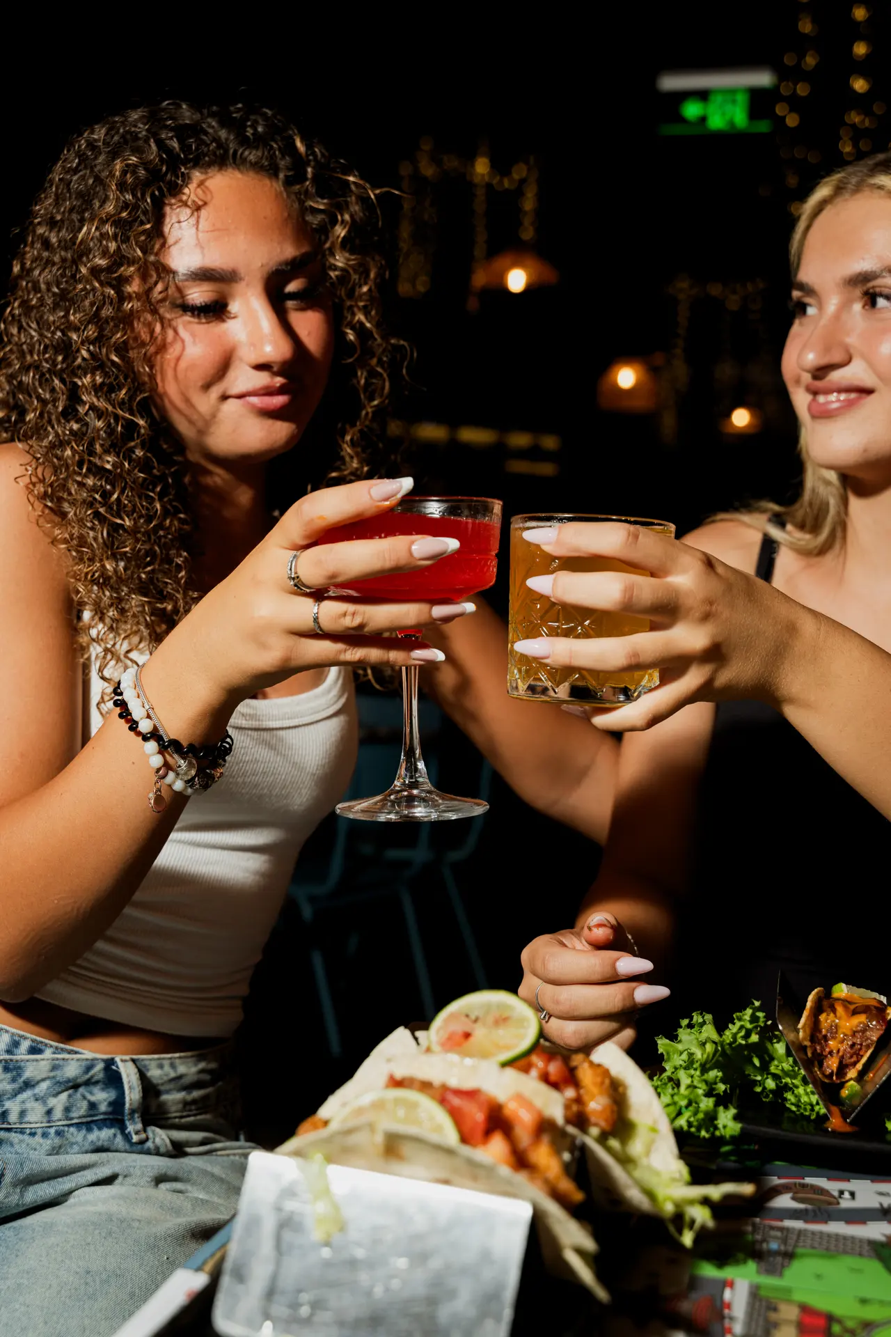 Due ragazze brindano al tavolo, con i tacos davanti